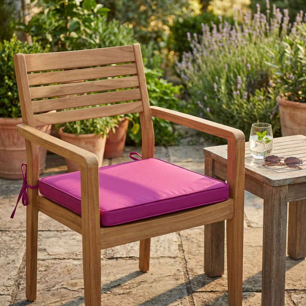 Dettaglio del cuscino in poliestere magenta su sedia in teak, con tavolino e bicchiere d'acqua.