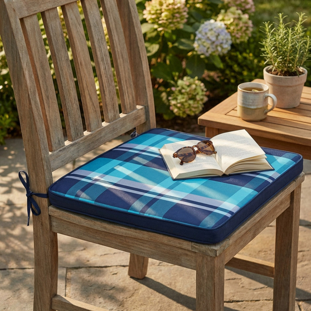 Cuscino Real Seduta Large a quadri blu posizionato su una sedia in legno da esterni con libro e occhiali da sole.