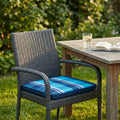 Cuscino Real Seduta blu a quadri posizionato su una sedia in rattan intrecciato in un giardino rigoglioso.