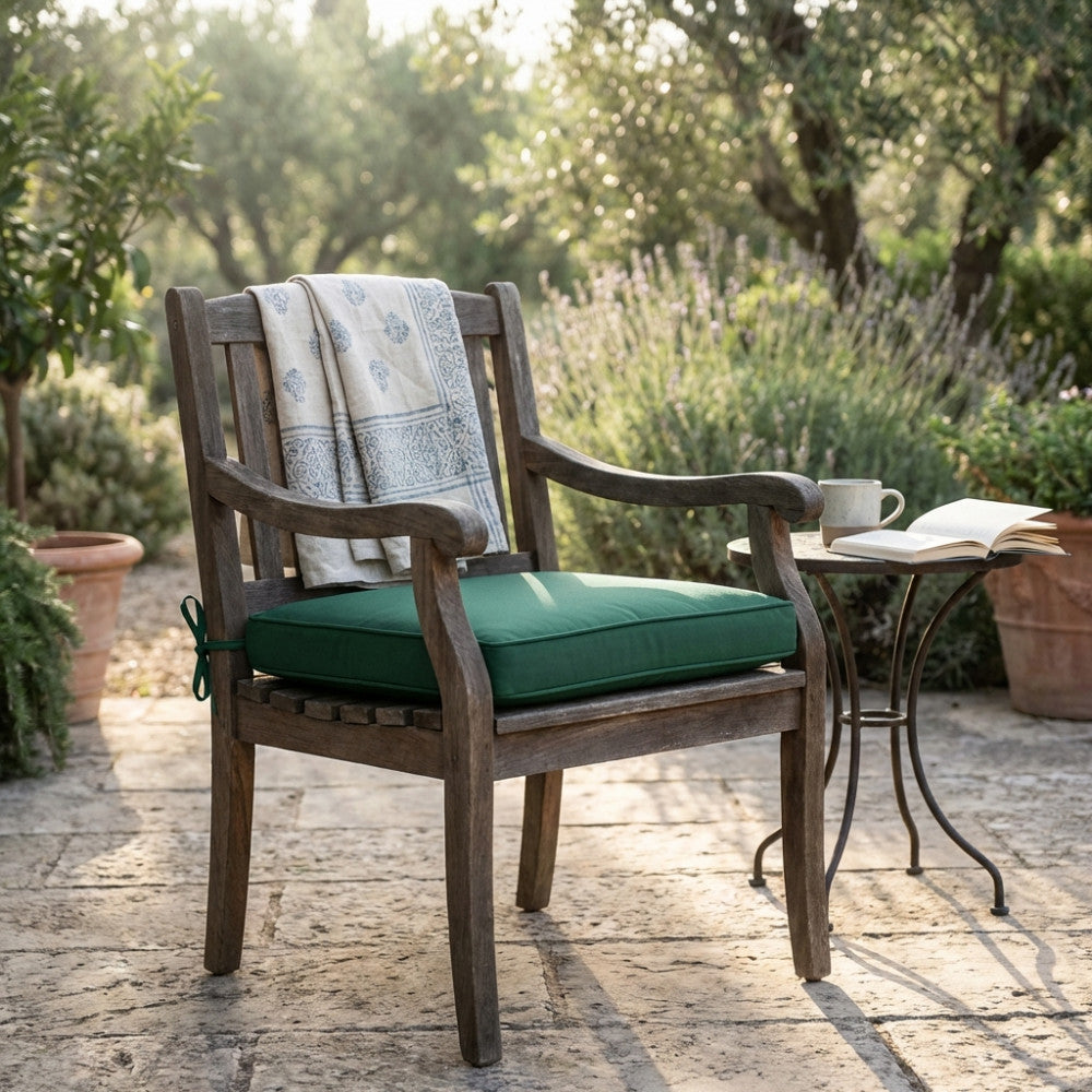 Cuscino verde scuro su una sedia in legno in un giardino, accompagnato da un tavolino in metallo con una tazza e un libro aperto.