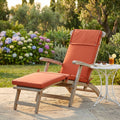 Cuscino Real Steamer arancione posizionato su una sdraio in legno in un rigoglioso giardino.