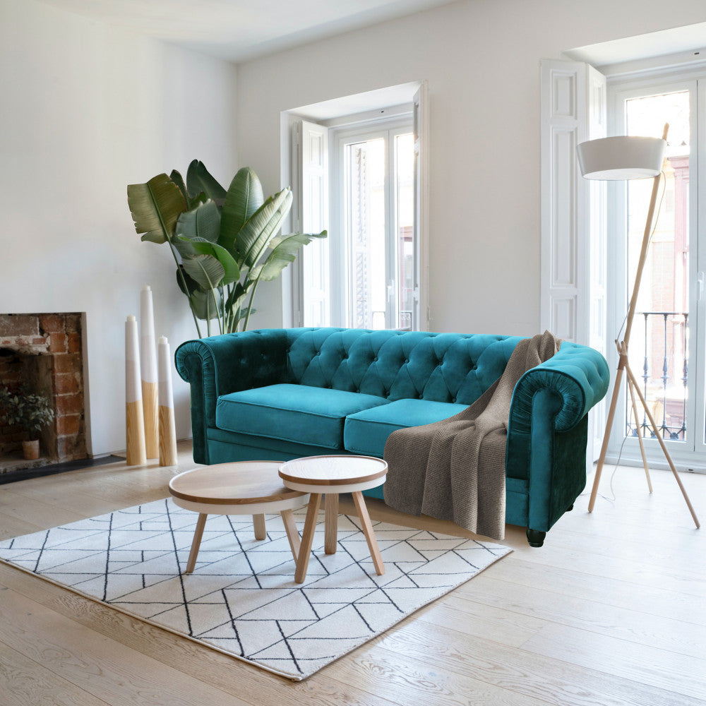 Divano Chesterfield in velluto blu in un salotto moderno, abbinato a tavolini in legno chiaro e tappeto geometrico.