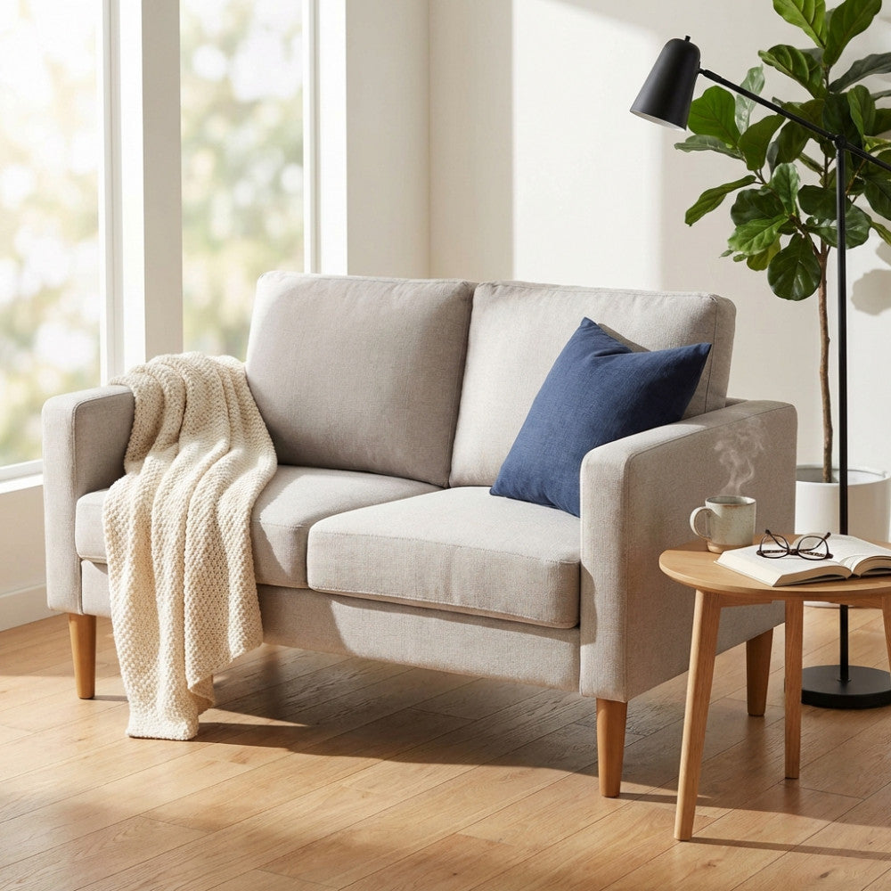 Divano 2 posti Ramsey in tessuto beige ambientato in un salotto luminoso con coperta e cuscino blu.