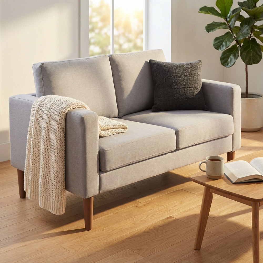 Divano 2 posti Ramsey in tessuto grigio chiaro, ambientato in un luminoso soggiorno con cuscino scuro e coperta in maglia.