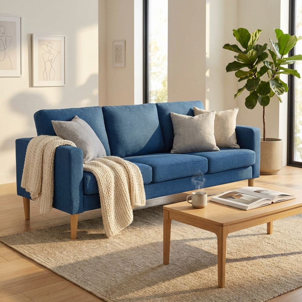 Divano 3 posti Ramsey in tessuto blu, ambientato in un soggiorno luminoso con tavolino in legno e pianta decorativa.