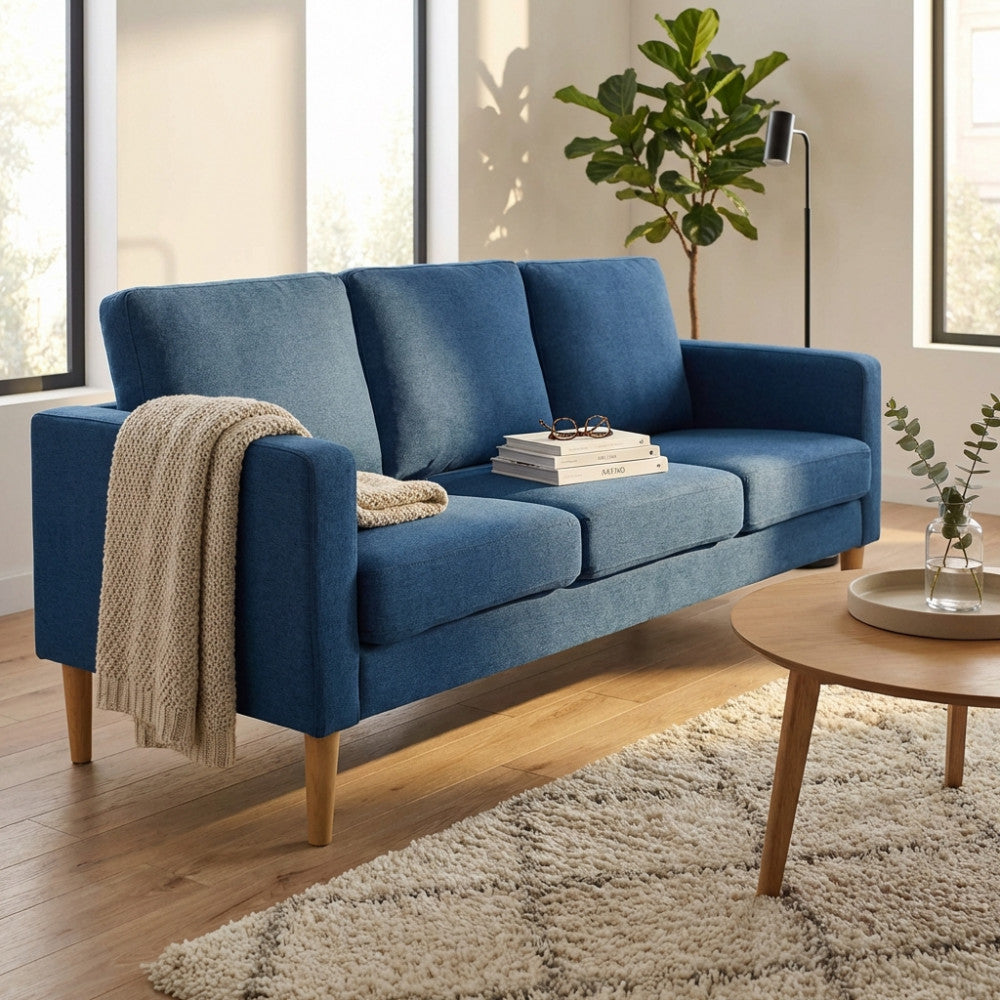 Vista angolare del divano Ramsey blu con cuscini decorativi e coperta in maglia, illuminato da calda luce naturale.