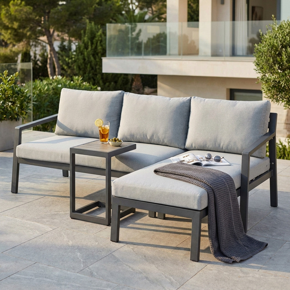 Divano Komodo ambientato su una terrazza soleggiata, con un drink rinfrescante sul tavolino integrato e una coperta grigia adagiata sulla chaise longue.
