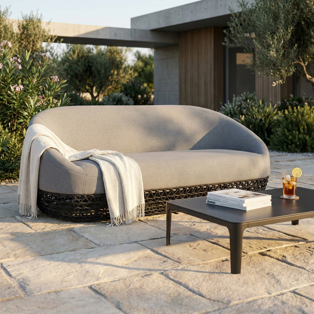 Divano moderno Lafayette grigio con base in corda intrecciata nera, ambientato in un patio esterno di lusso con tavolino scuro.