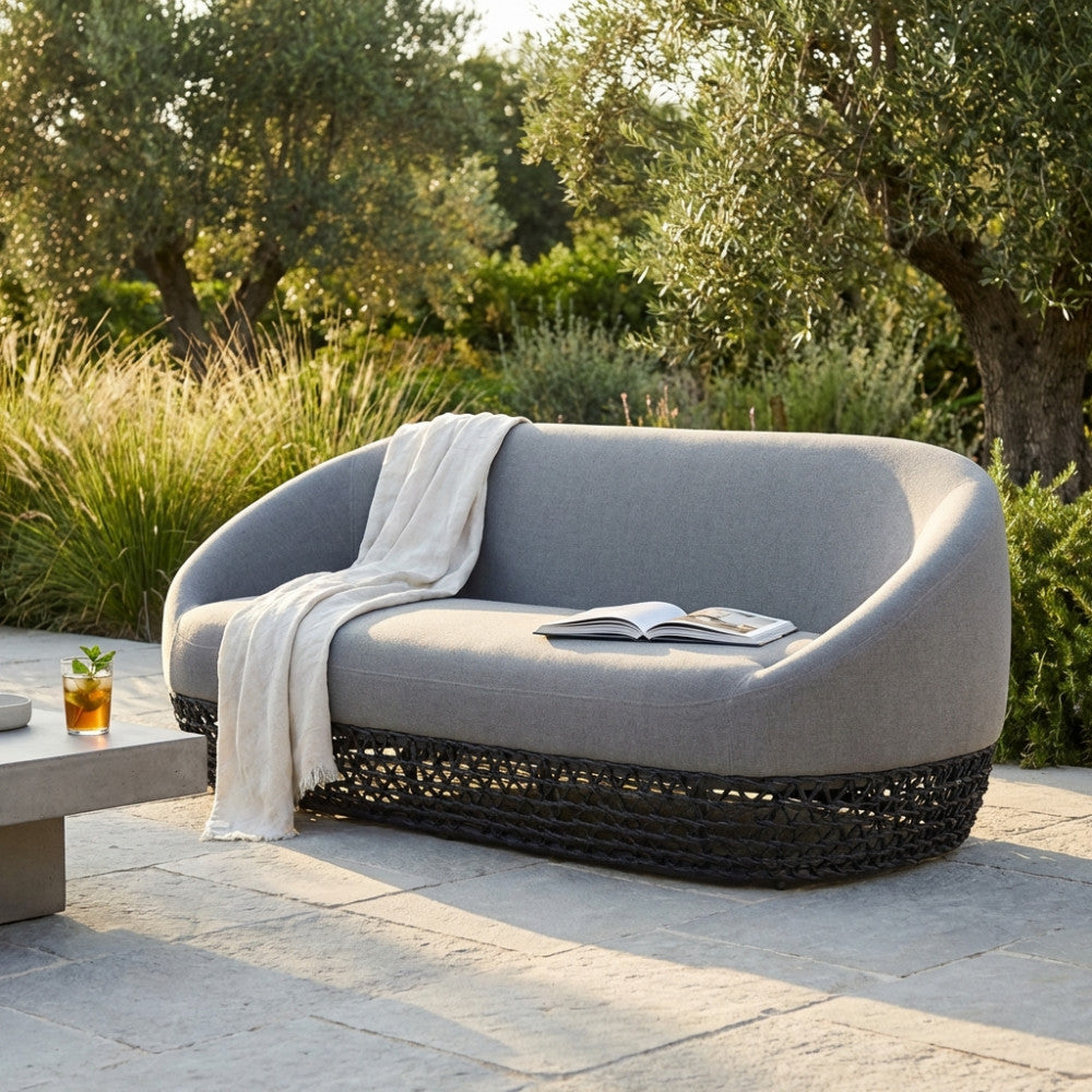 Divano da esterno Lafayette in tessuto grigio e alluminio, decorato con un plaid chiaro e un libro, circondato da vegetazione mediterranea.