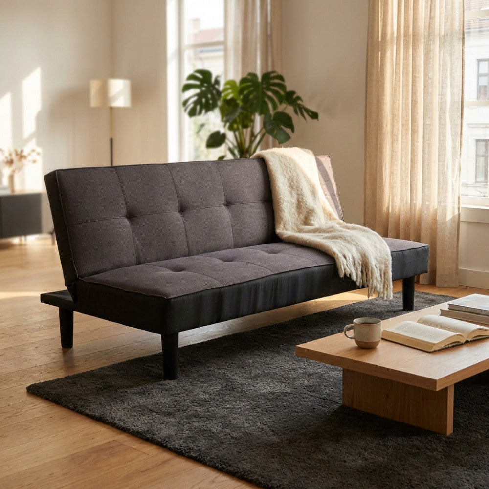 Vista frontale del divano letto Alvin color antracite in un salotto moderno, decorato con un plaid chiaro e cuscini.