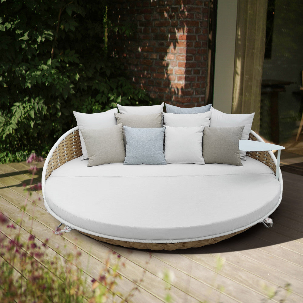 Divano letto rotondo Formosa posizionato a terra su un patio in legno, allestito con molteplici cuscini decorativi per un comfort ottimale.