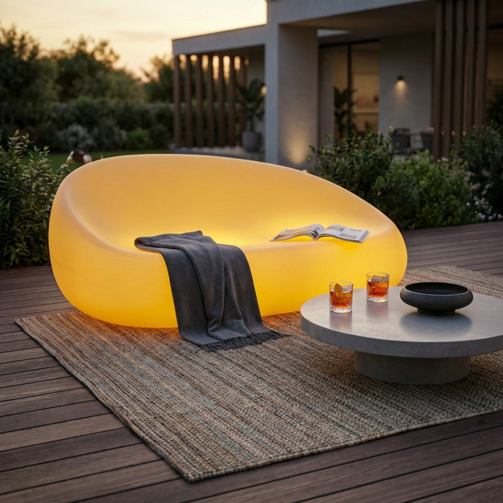 Divano luminoso Lepus su una terrazza in legno con una coperta appoggiata, vicino a un tavolino rotondo in cemento.