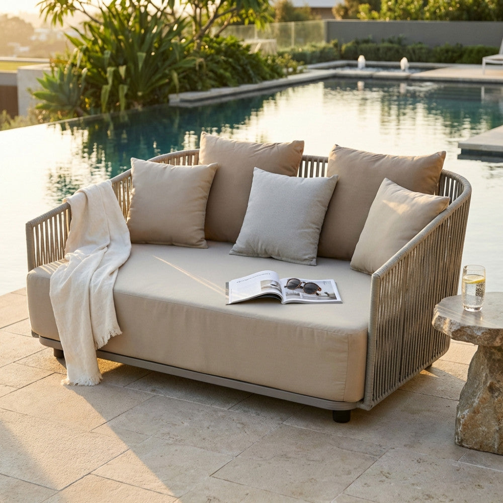 Divano Mendoza a due posti in poliestere beige, posizionato a bordo piscina al tramonto con cuscini decorativi.