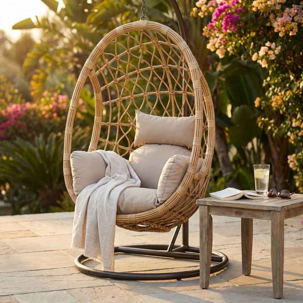 Dondolo sospeso in rattan sintetico intrecciato su struttura in acciaio marrone, ambientato in un rigoglioso giardino al tramonto con cuscini beige.