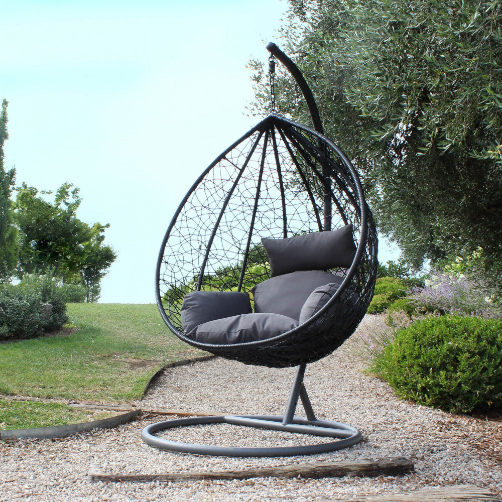 Dondolo da giardino in wicker nero posizionato su ghiaia con ulivi sullo sfondo.