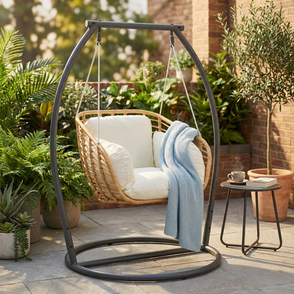 Poltrona sospesa Glasgow ambientata in un rigoglioso giardino con cuscini bianchi e coperta azzurra.