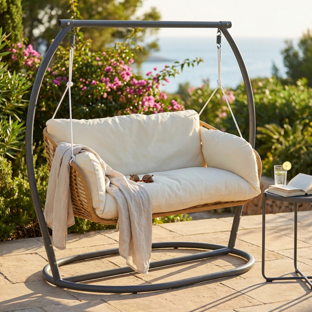 Dondolo Glasgow 2 posti in metallo beige ambientato in un giardino soleggiato con vista mare.