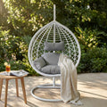 Dondolo Panarea in wicker bianco ambientato in un giardino lussureggiante, con cuscini grigi e una coperta in lino.