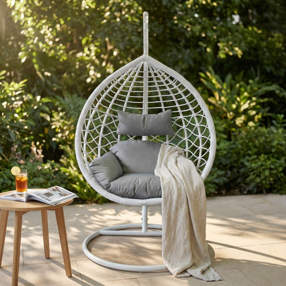 Dondolo Panarea in wicker bianco ambientato in un giardino lussureggiante, con cuscini grigi e una coperta in lino.