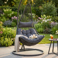 Dondolo sospeso Ponza in wicker nero marrone ambientato in un giardino rigoglioso con cuscini grigi.