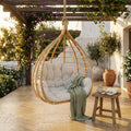Dondolo sospeso Calypso in bamboo marrone chiaro con cuscino bianco, ambientato in un patio lussuoso al tramonto.
