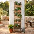 Fioriera Chelsey in legno riciclato marrone, allestita con piante aromatiche e fiori, appoggiata a un pilastro bianco in un patio esterno.