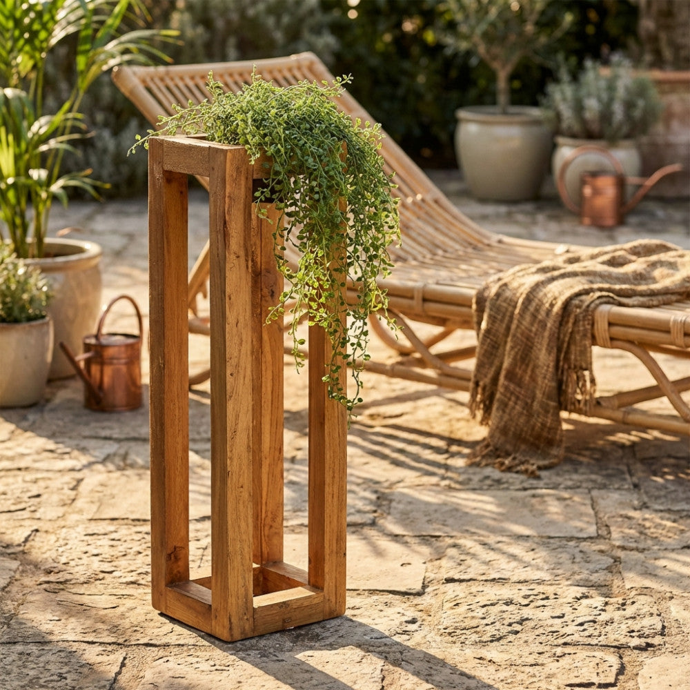 Fioriera Cecily in legno marrone allestita in un patio esterno in pietra, affiancata da un lettino in bambù e una pianta ricadente.
