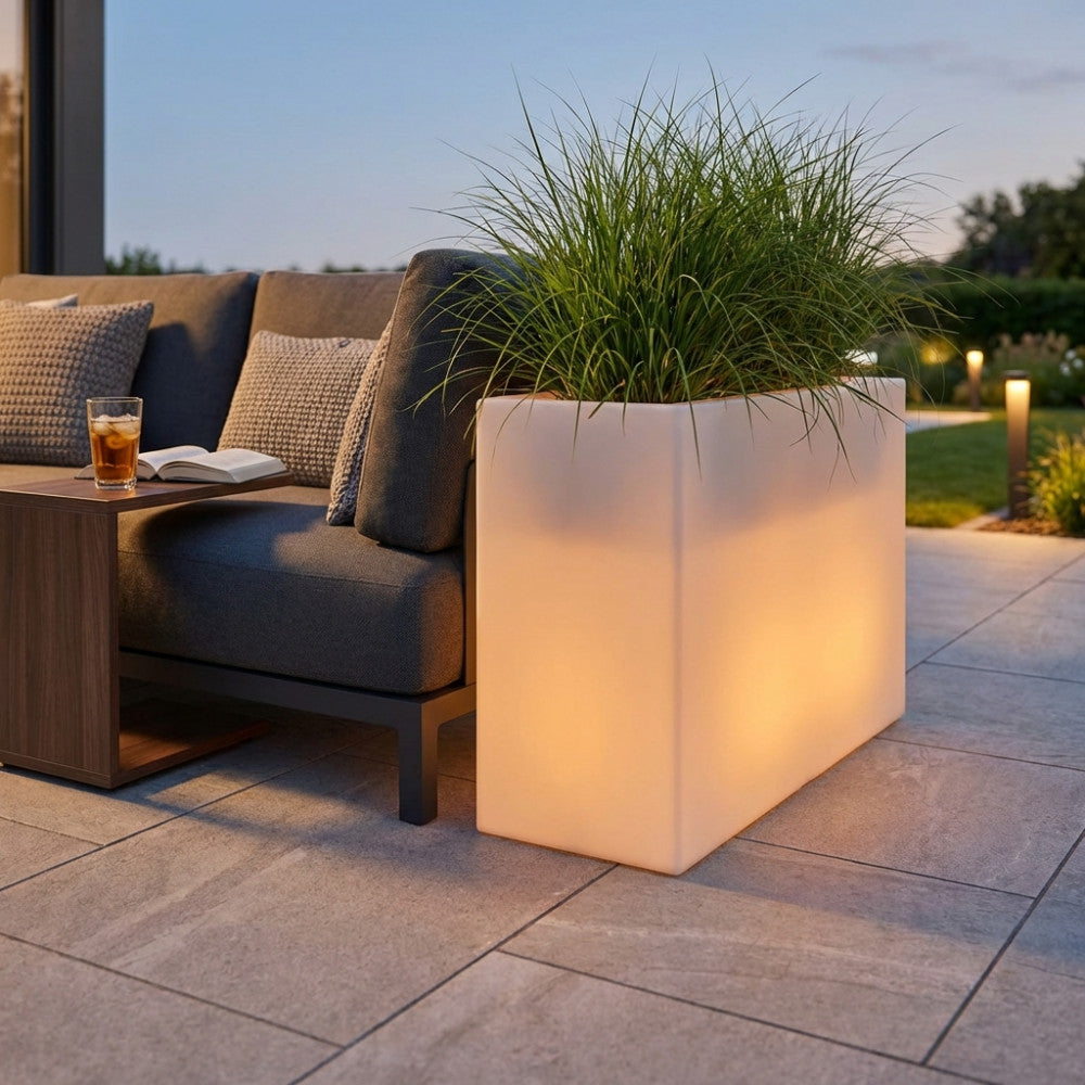 Fioriera luminosa Sculptor 50 in polipropilene bianco, accesa con luce calda, posizionata su un patio moderno accanto a un divano grigio.
