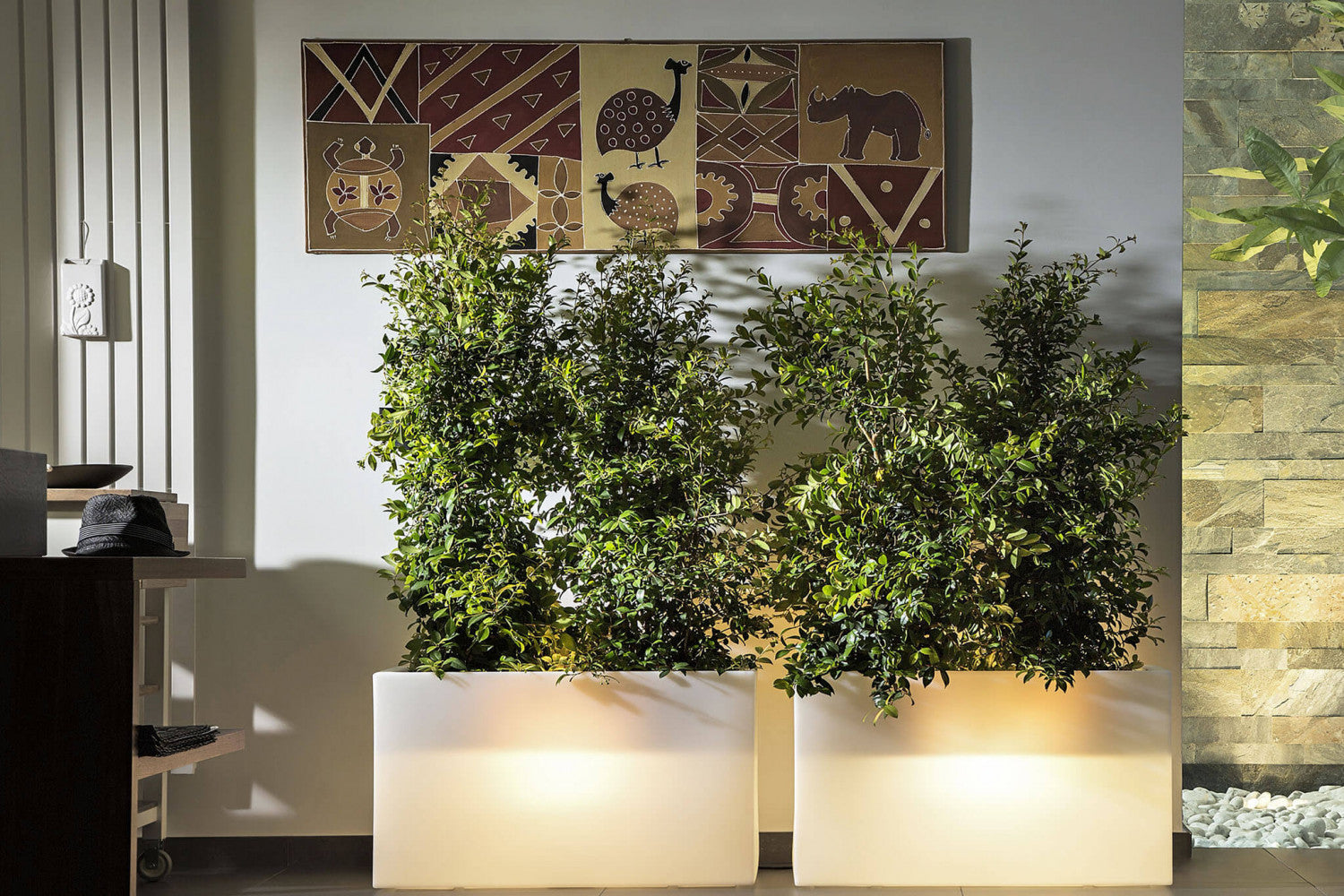 Coppia di fioriere luminose in polipropilene bianco con piante alte verdi, collocate in un ambiente interno con arte etnica a parete.