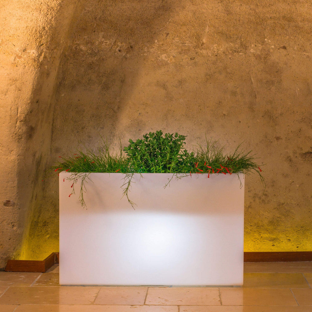 Fioriera luminosa in polipropilene bianco con piante ricadenti, ambientata in una suggestiva alcova in pietra rustica.
