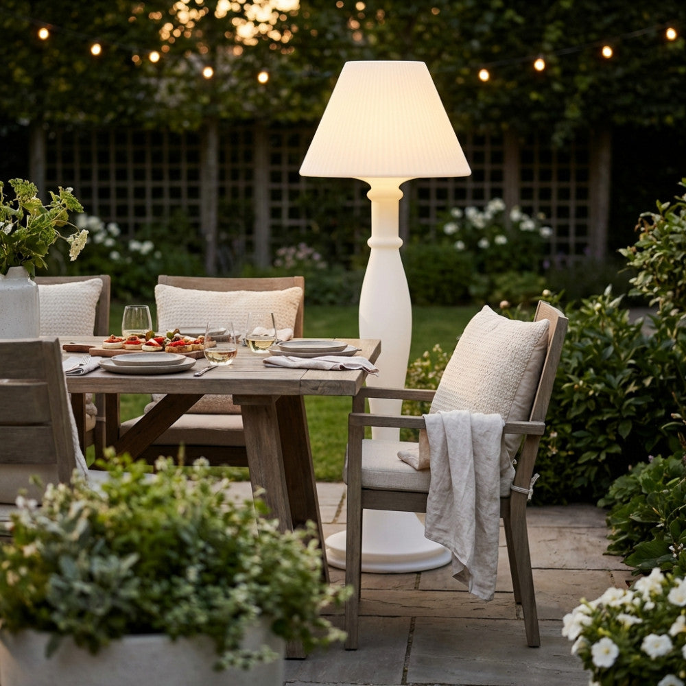 Dettaglio della lampada da terra Aries accanto a un tavolo in legno con sedie da giardino e luci a corda.