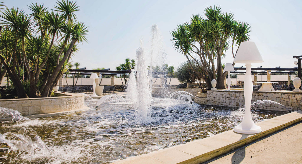 Lampada da terra Aries posizionata a bordo piscina vicino a una grande fontana e palme lussureggianti.