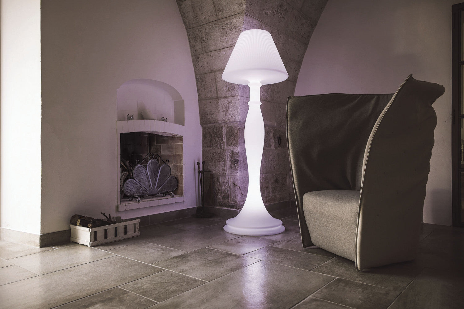 Lampada da terra Aries che diffonde una luce soffusa in un interno rustico accanto a una poltrona moderna.