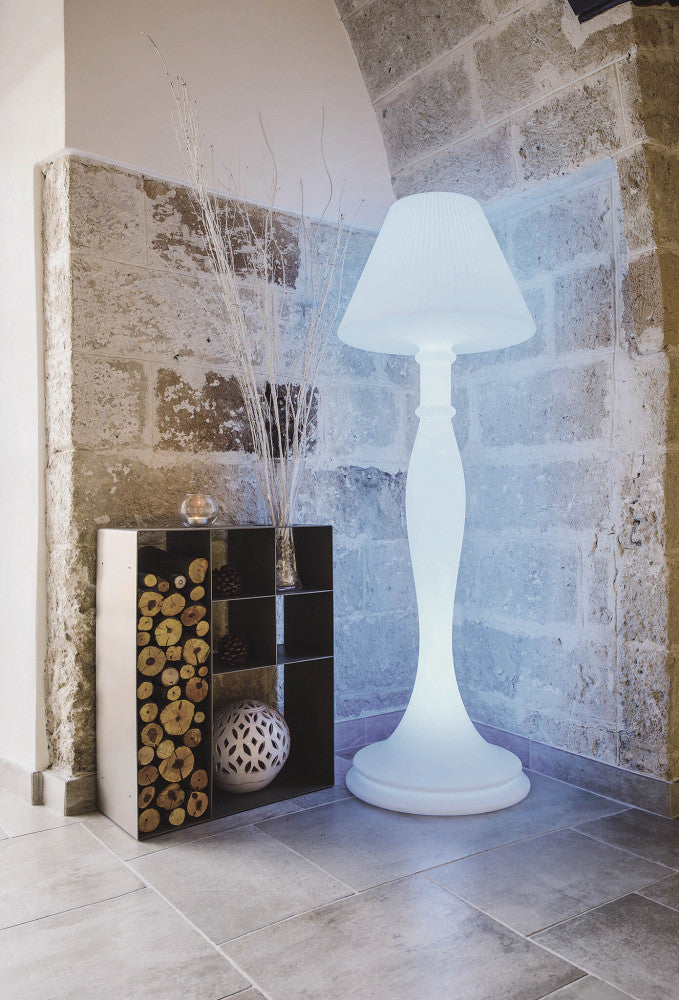 Lampada da terra Aries illuminata di bianco freddo vicino a una parete in pietra e un portalegna di design.