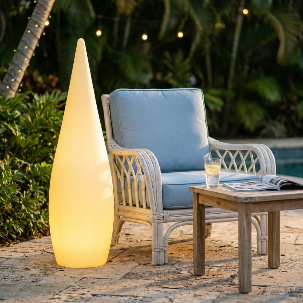 Lampada da terra a forma di goccia gialla illuminata posizionata su un patio in pietra accanto a una poltrona in rattan.
