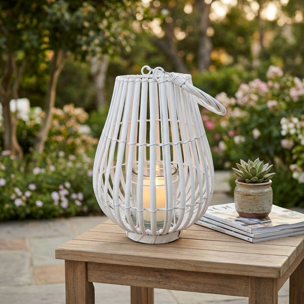 Lanterna Celeste in vimini bianco con candela accesa nel cilindro di vetro, posizionata su un tavolino in legno in un giardino fiorito.