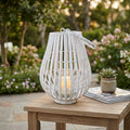 Lanterna Celeste in vimini bianco con candela accesa nel cilindro di vetro, posizionata su un tavolino in legno in un giardino fiorito.