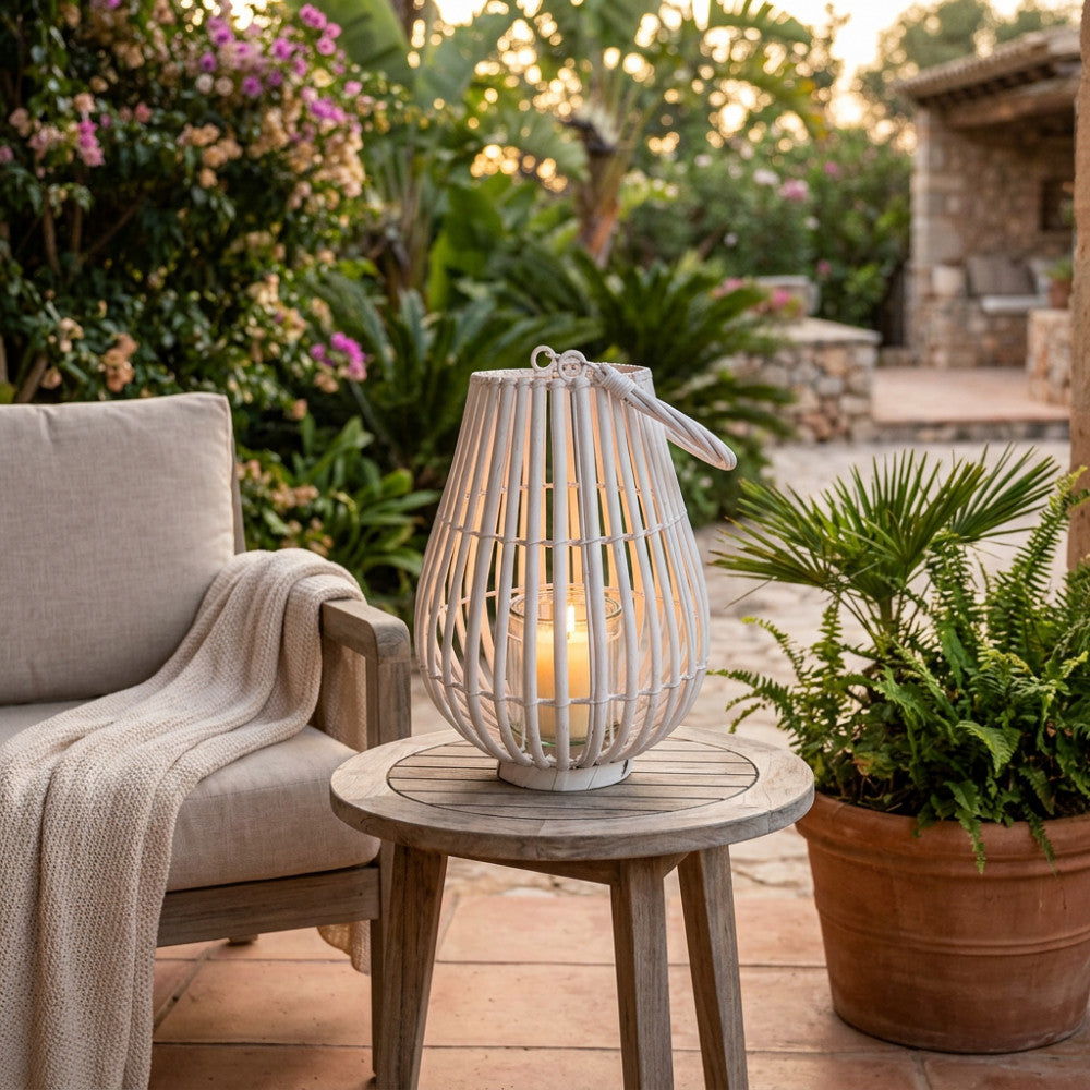 Lanterna decorativa in vimini e vetro su un tavolino rotondo in legno, accanto a una poltrona con cuscini chiari e piante rigogliose in un patio elegante.