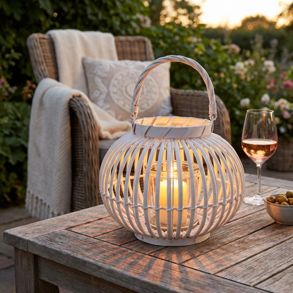 Lanterna in vimini bianco illuminata da una candela interna, situata su un tavolo in legno al tramonto, con un calice di vino e una poltrona in rattan sullo sfondo.