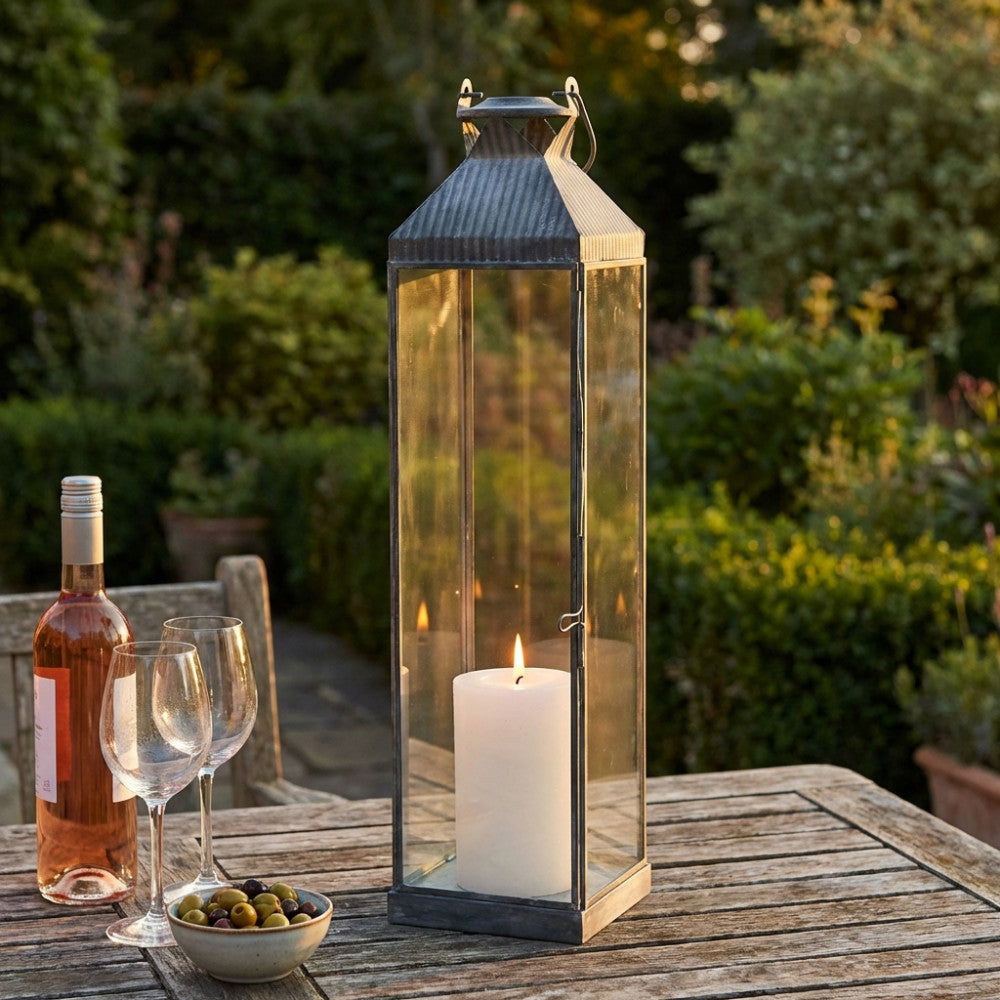 Lanterna Harmony Media in metallo grigio e vetro posizionata su un tavolo in legno da giardino al tramonto.