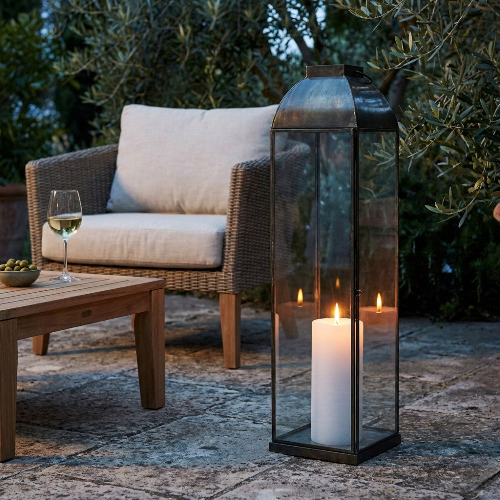 Alta lanterna Karrie Grande posizionata su un patio in pietra accanto a una poltrona intrecciata, illuminata da una grande candela.