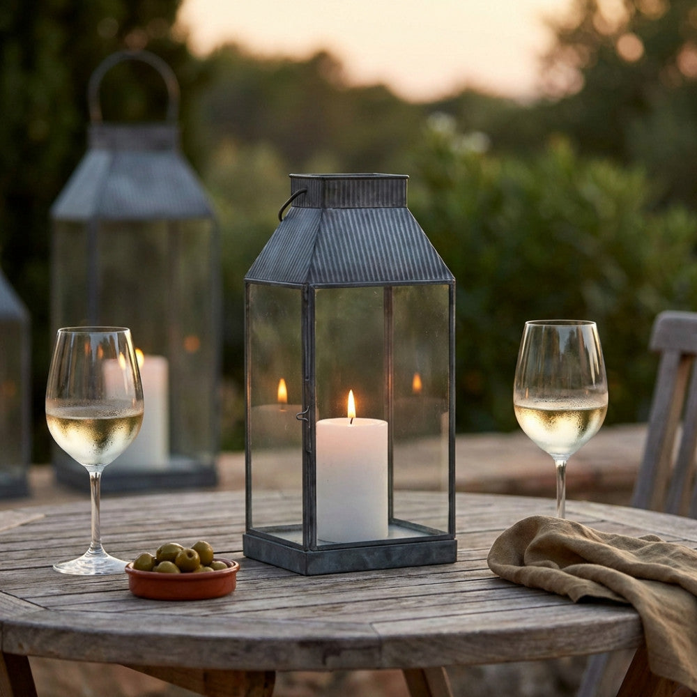 Lanterna Kassy Piccola in vetro e metallo grigio su un tavolo in legno all'aperto, illuminata da una candela, con due calici di vino bianco.
