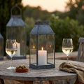 Lanterna Kassy Piccola in vetro e metallo grigio su un tavolo in legno all'aperto, illuminata da una candela, con due calici di vino bianco.