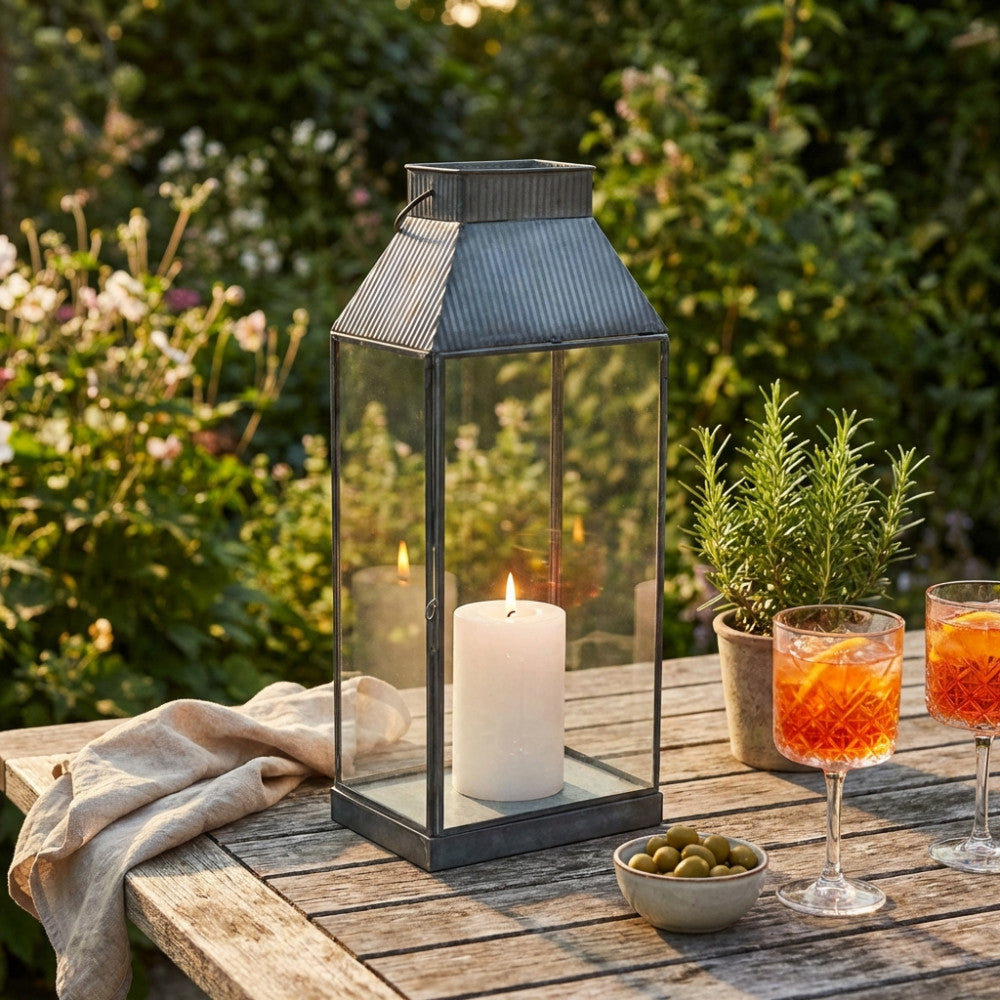Lanterna Kassy Piccola accesa su un tavolo da giardino in legno, affiancata da cocktail e una pianta aromatica al tramonto.