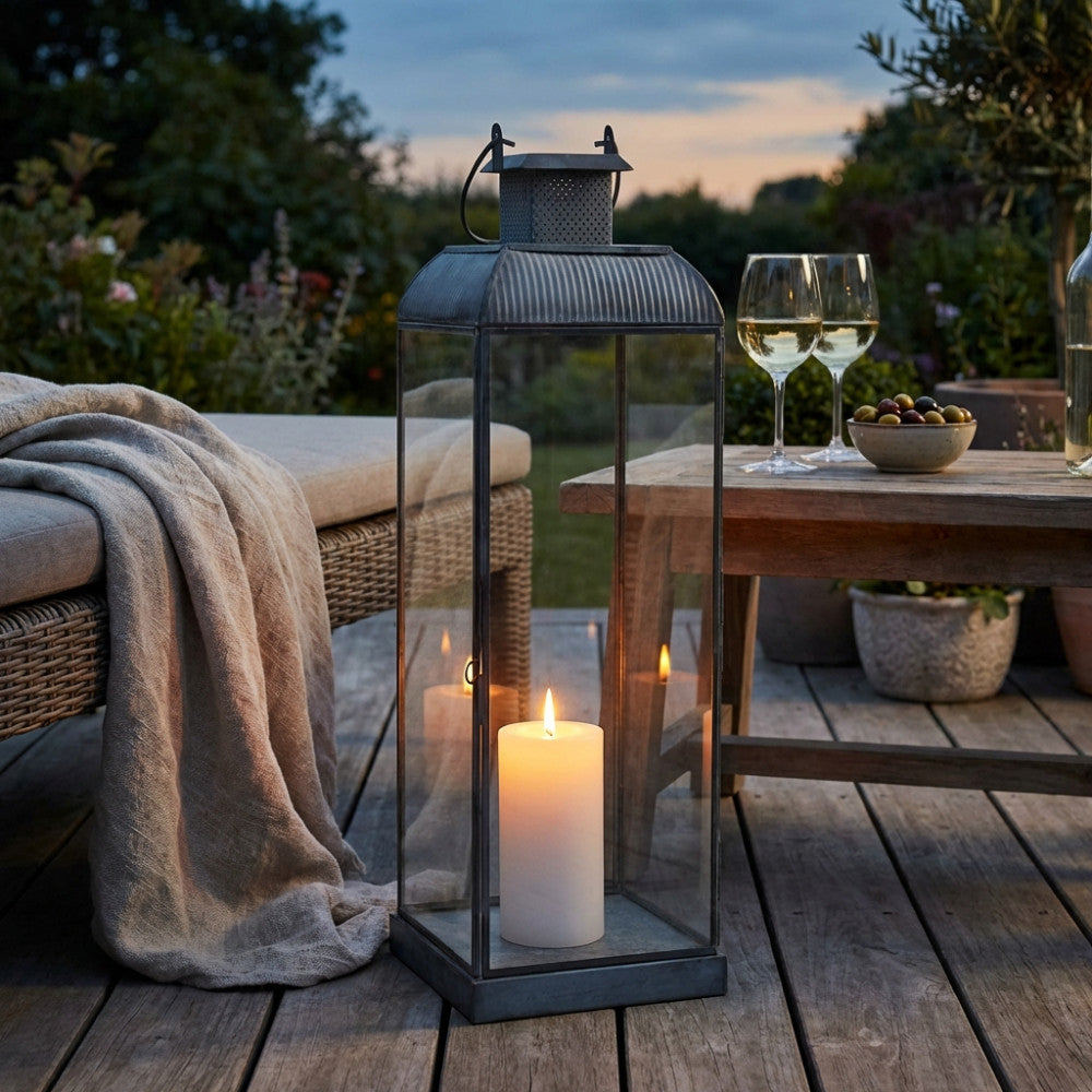 Lanterna decorativa illuminata da una candela, situata su un pavimento in legno all'aperto al tramonto, accanto a un divano in rattan e bicchieri di vino.