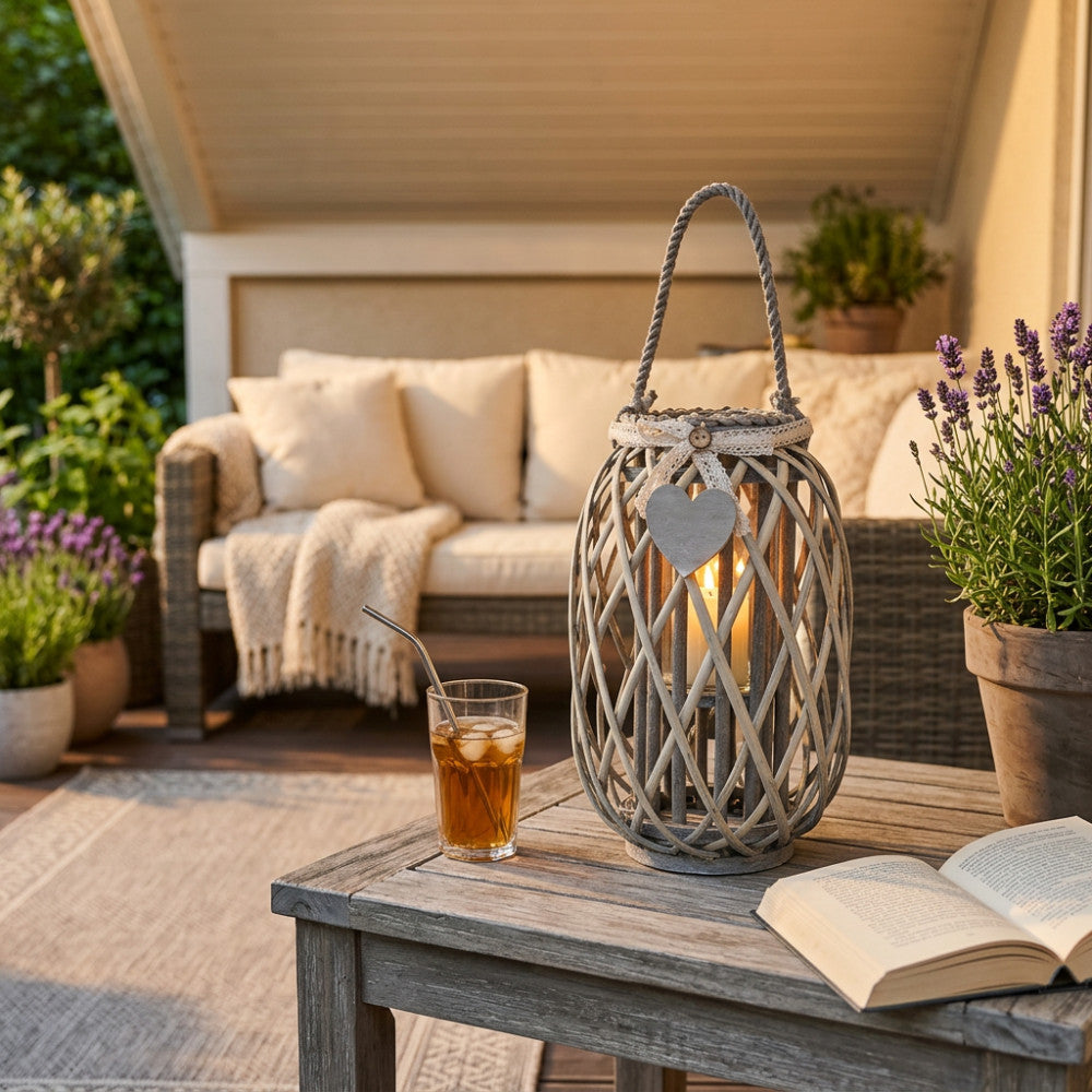 Lanterna Lison in vimini grigio con cuore decorativo, posizionata su un tavolino in legno grezzo in una veranda elegante, illuminata dalla luce calda del tramonto.