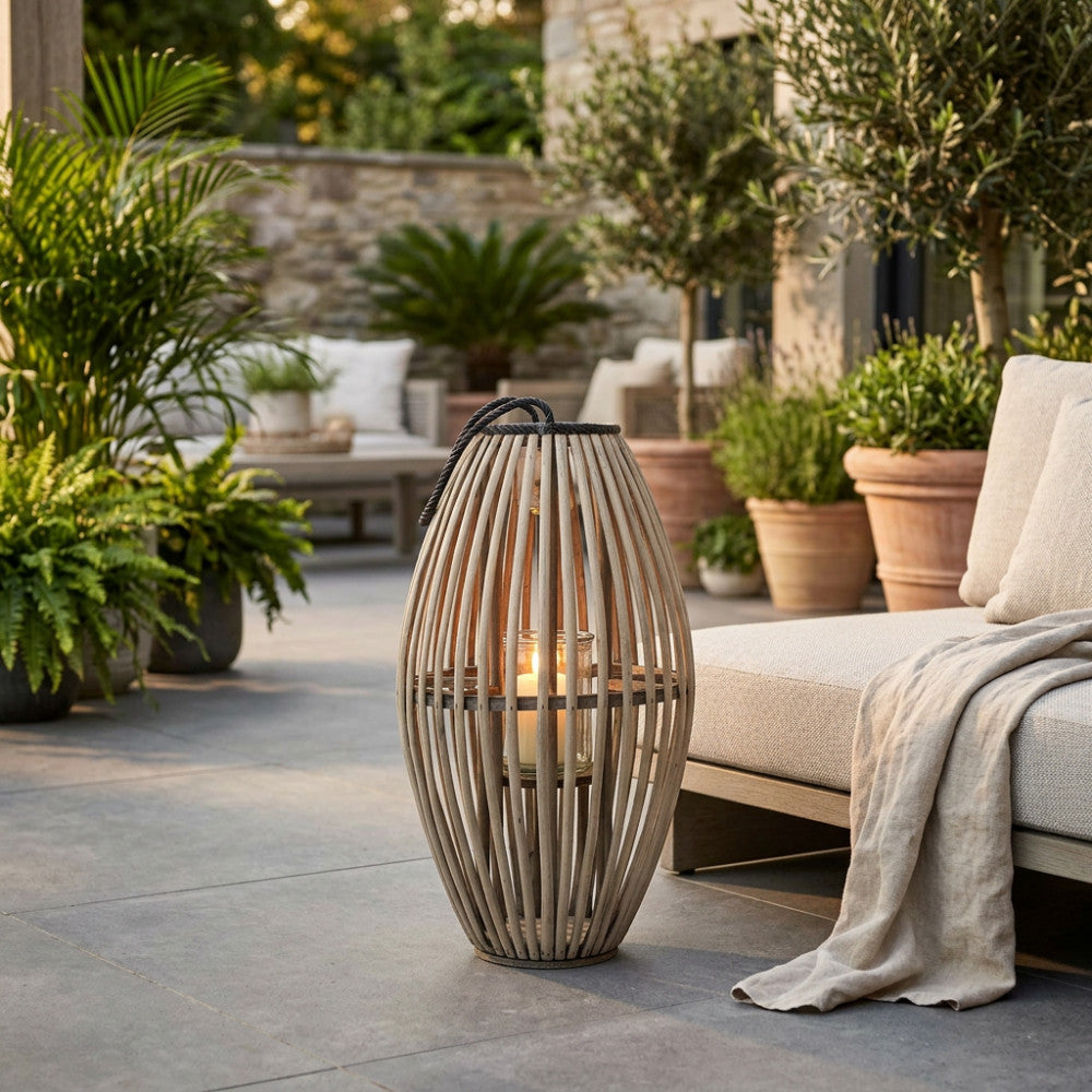 Lanterna Lucille in vimini grigio posizionata su una pavimentazione in pietra in un elegante patio esterno, accanto a un divano con cuscini chiari.