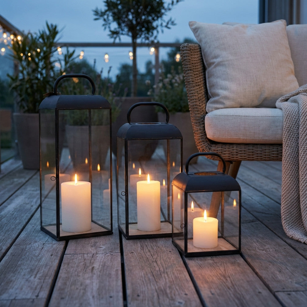 Lanterne di design in metallo scuro appoggiate su un pavimento a doghe in legno, creando un'atmosfera serale intima accanto a una poltrona intrecciata.
