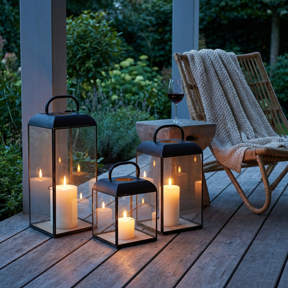 Set di tre lanterne antracite illuminate su un pavimento in legno a doghe in un patio esterno, accanto a una sedia in rattan con coperta intrecciata.
