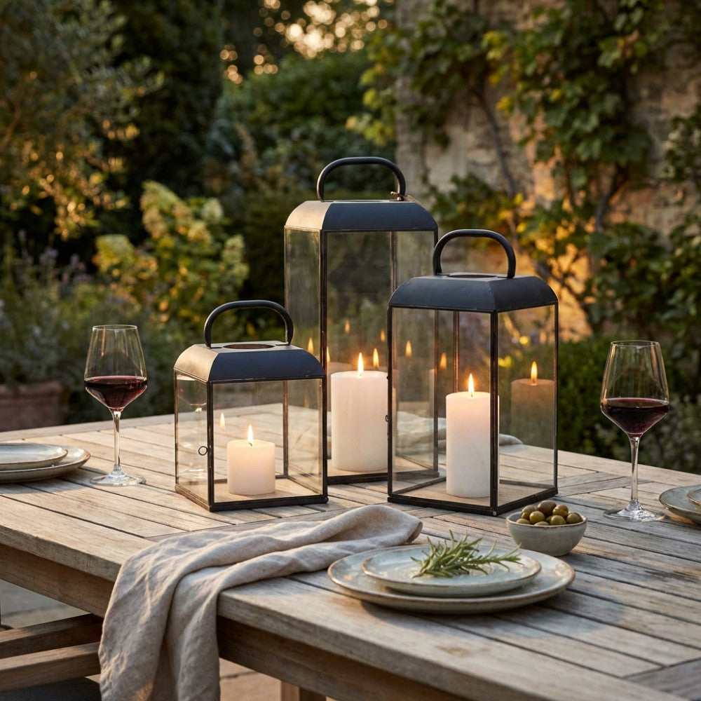 Dettaglio di lanterne moderne in metallo scuro e vetro su un tavolo in legno apparecchiato con calici di vino rosso, creando un'atmosfera serale elegante.